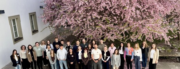 Gruppenfoto der neuen Nachwuchskräfte der Regierung der Oberpfalz beim Kennenlerntag im April 2026. Die Teilnehmenden stehen im Innenhof der Regierung vor einem großen rosa blühenden Baum.