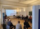 Wie gelingt gute Kommunikation im Klimaschutz? Beim Workshop der Regierung der Oberpfalz entwickelten die Teilnehmenden gemeinsam neue Ansätze für Gespräche vor Ort. Seminarteilnehmende in Stuhlkreis schauen auf eine Präsentation, Vortrag zum Gesprächsabschluss im Klimaschutz.