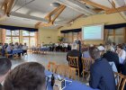 Veranstaltungssaal mit mehreren runden Tischen, festlich gedeckt. Im Hintergrund spricht ein Vertreter der Regierung der Oberpfalz am Rednerpult, rechts ist eine Leinwand mit dem Titel der Freisprechungsfeier zu sehen. Eine Blaskapelle begleitet die Veranstaltung musikalisch.