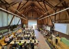 Blick in eine große, holzverkleidete Halle mit festlich gedeckten Tischen und zahlreichen Gästen. Auf der Bühne rechts spricht ein Redner bei der Freisprechungsfeier im Ausbildungsberuf „Landwirt“, während eine Blaskapelle auf der Bühne sitzt. 