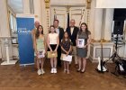 Drei Schülerinnen der Mittelschule Lam stehen mit Urkunden und Preisumschlägen gemeinsam mit Lehrkräften und Ehrengästen vor dem blauen Banner der Regierung.