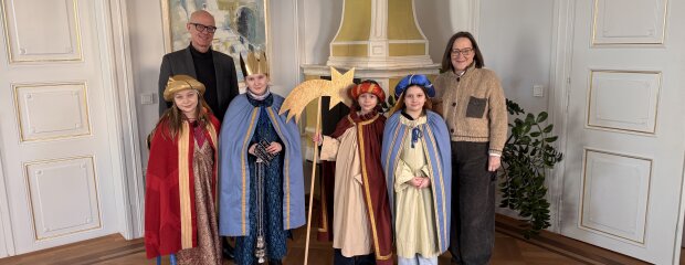 Regierungspräsident Walter Jonas und Regierungsvizepräsidentin Christiane Zürn stehen gemeinsam mit vier als Sternsinger verkleideten Kindern in einem historischen Raum der Regierung der Oberpfalz. Die Kinder tragen bunte Gewänder, Kronen und einen Stern.